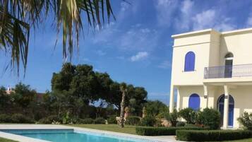 Blick auf einen Pool vor einem zweistöckigen Gebäude mit Balkon, umgeben von Grün und blauem Himmel.