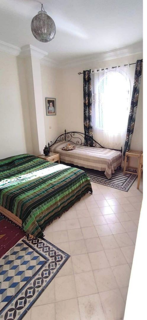 Un dormitorio con cama, mesita de noche, lámpara, ventana con cortinas y alfombra estampada.