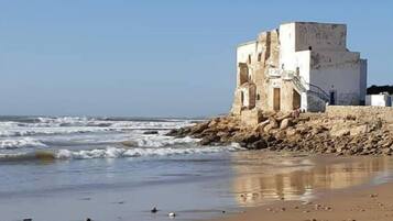 Ein Küstengebäude mit steinernem Sockel und weißer Oberstruktur, gelegen an einem Sandstrand mit vereinzelten Felsen.