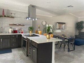 Una cocina moderna con armarios oscuros, encimera blanca y una isla central. Hay un comedor con mesa y sillas, y una sala de estar con sofá y una planta.