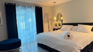 A hotel room with a large bed, dark curtains, a round ottoman, a mirror, and a potted plant.