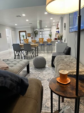Una sala de estar moderna con sofá, reposapiés y una mesita de centro. Hay un comedor con sillas y una mesa, y una cocina al fondo.