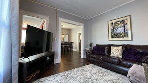 Una sala de estar con una televisión de pantalla plana sobre un mueble bajo, un sofá de cuero marrón con almohadones decorativos, una alfombra estampada y un cuadro en la pared.