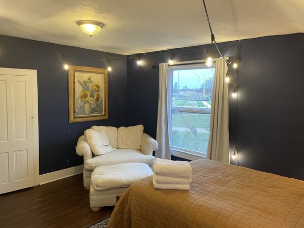 A bedroom with a bed, a white armchair, a painting, and a window with curtains. - Home in Berrien Springs (Berrien Springs)