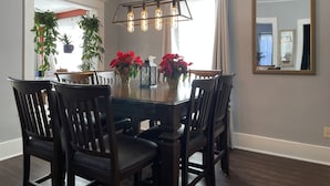 Comedor con mesa de madera oscura, seis sillas y una araña con bombillas expuestas. Hay plantas en macetas y flores rojas sobre la mesa.