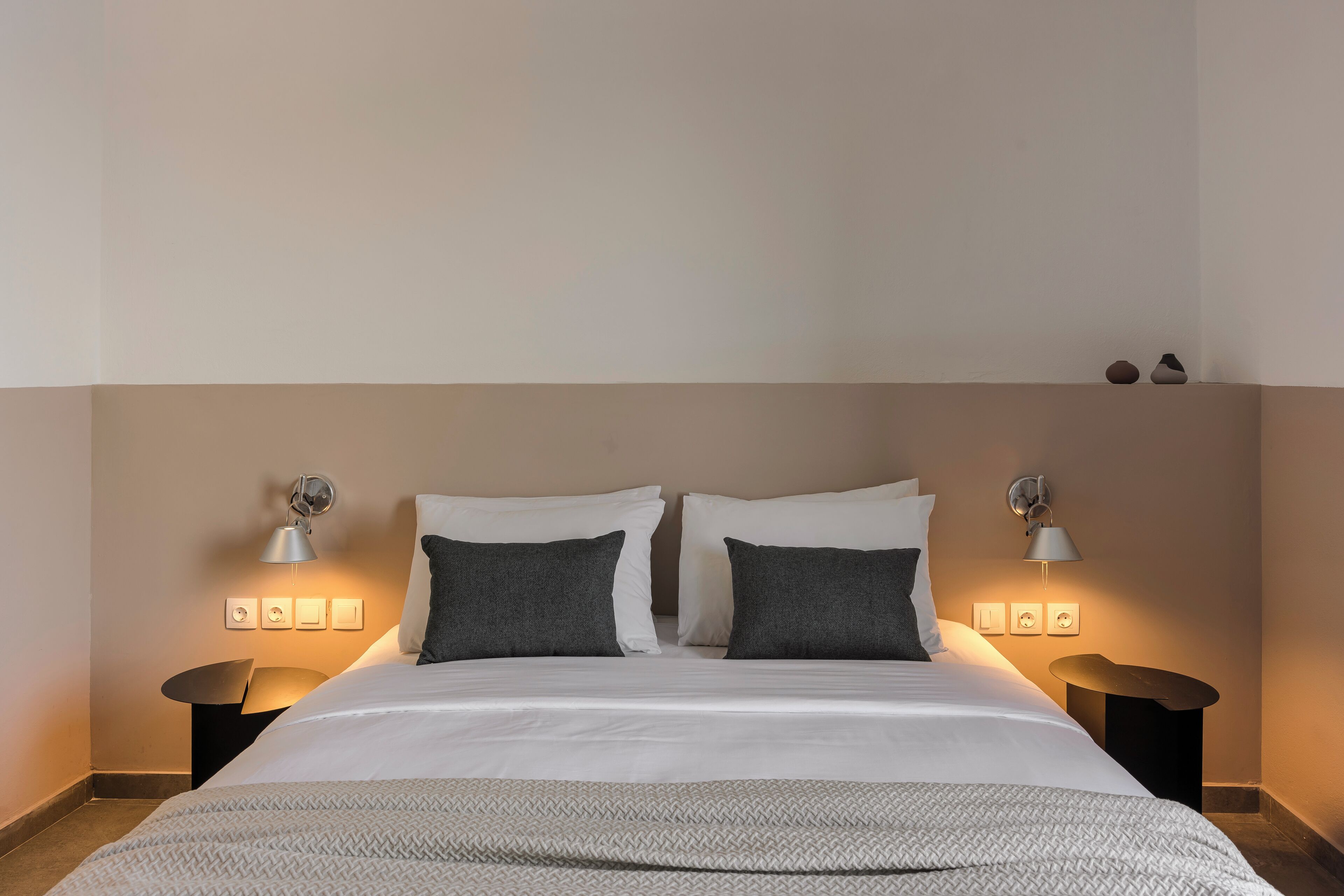 Un lit bien fait, recouvert d’une couvre-lit à motifs blancs et gris, est flanqué de deux tables de chevet surmontées de lampes. Le tout est placé devant un mur uni, au-dessus duquel se trouve une étagère.
