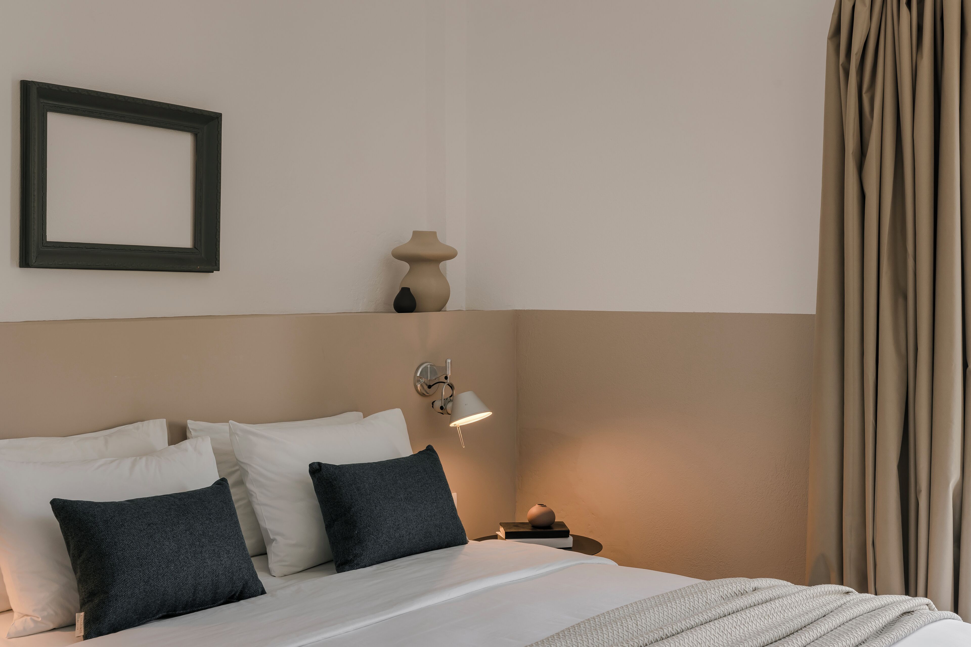 Una cama bien hecha con almohadas blancas y gris oscuro, una mesita de noche con una lámpara y un marco de fotos colgado en la pared.