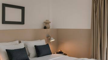 Una cama bien hecha con almohadas blancas y gris oscuro, una mesita de noche con una lámpara y un marco de fotos colgado en la pared.