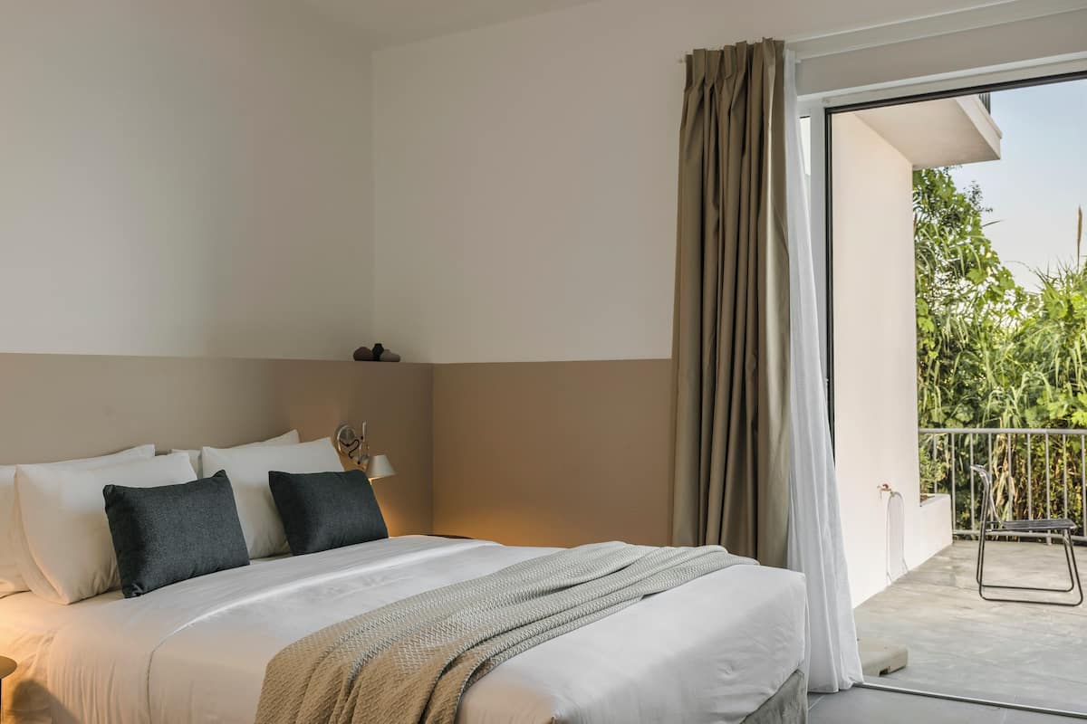 A hotel room with a bed, a nightstand, a lamp, and a view of a balcony with plants.
