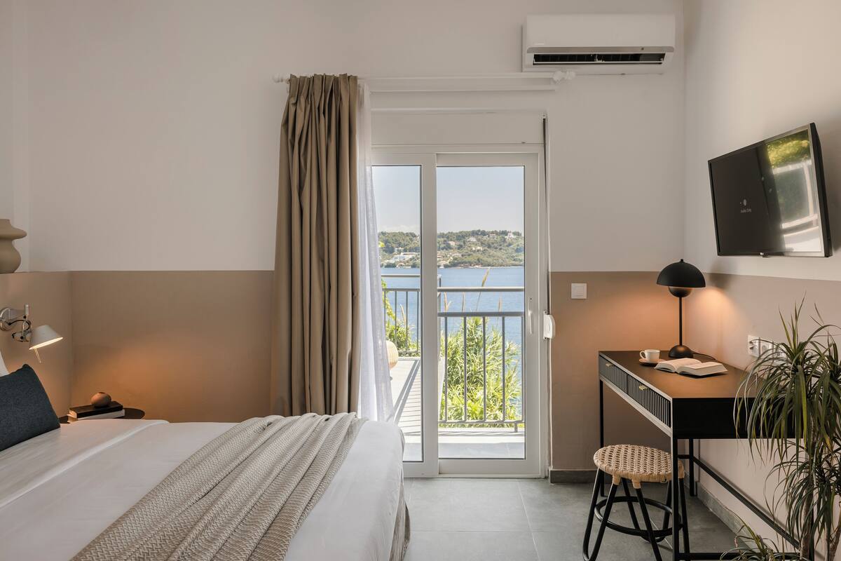 A hotel room with a bed, a desk with a chair, a TV, and a balcony with a view of the sea.