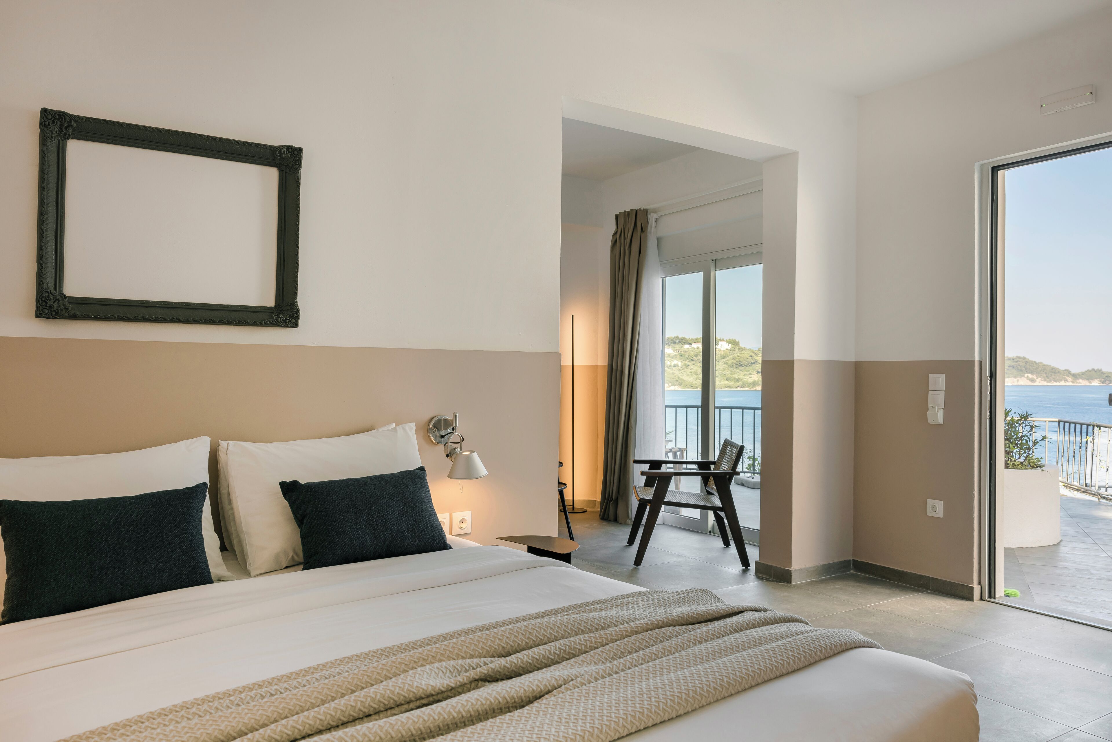 A hotel room with a large bed, a chair, a balcony with a view of the sea, and a framed picture on the wall.