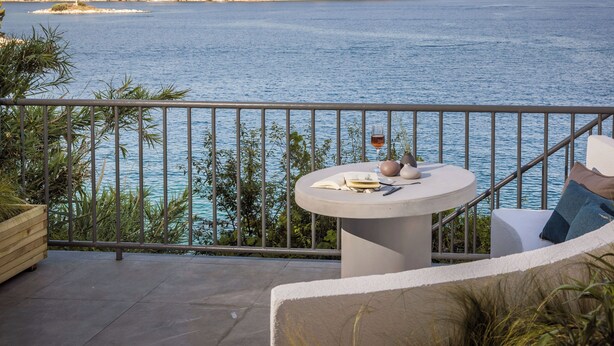 Un balcone con vista sul mare, un tavolo tondo con un libro e un bicchiere, e una sedia con un cuscino.