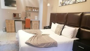 A neatly made bed with a brown blanket, a dark brown headboard, and white pillows. There is a small desk with a chair and a microwave above it. - On Hill Lodge (Bloemfontein)