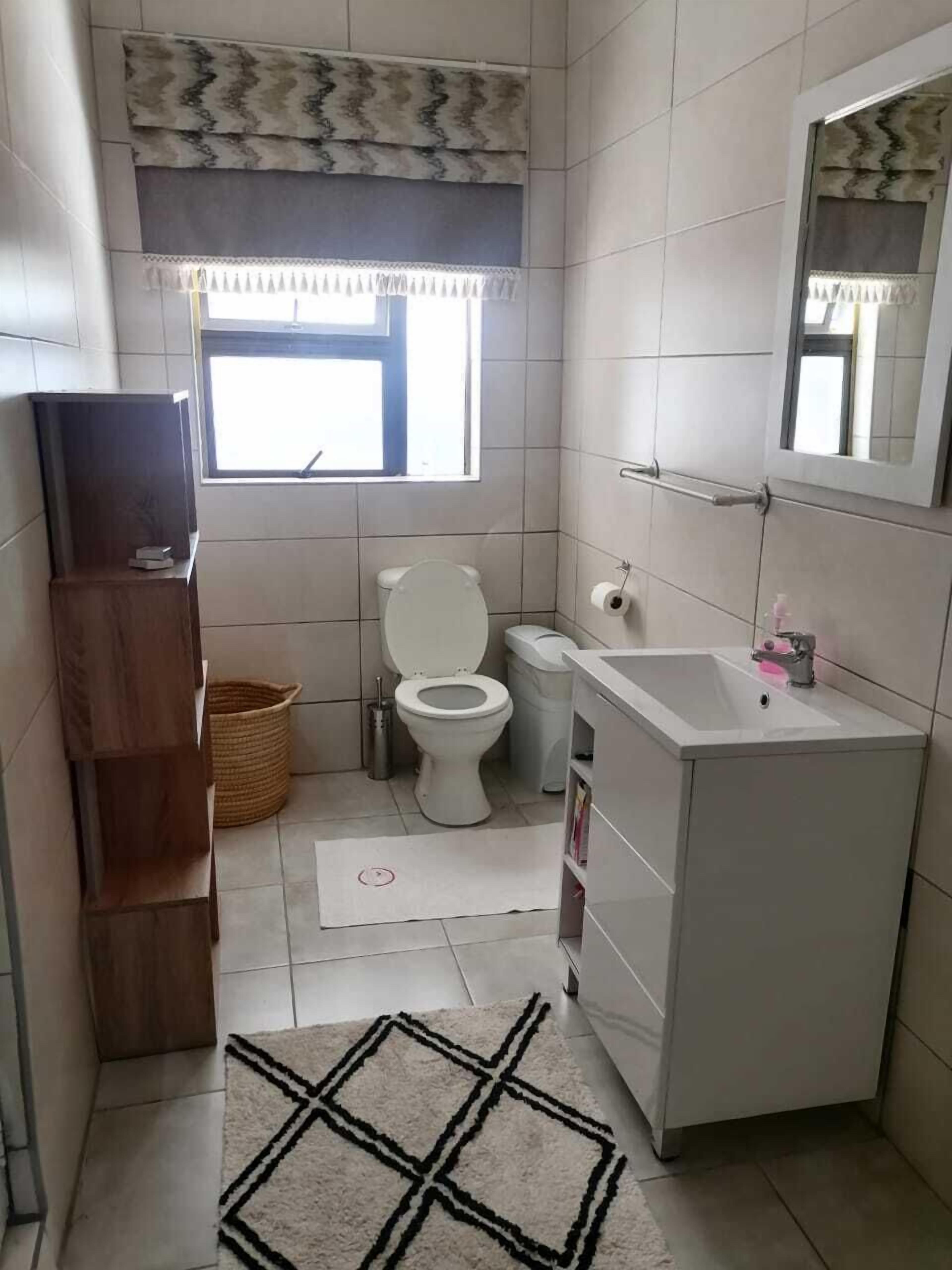 A small, clean bathroom with a toilet, sink, and mirror. There is a window above the toilet and a wooden shelf to the left.