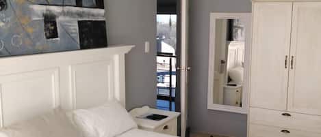 Un dormitorio con cama, cortinas blancas, un cuadro, un armario y vistas a través de una puerta abierta.
