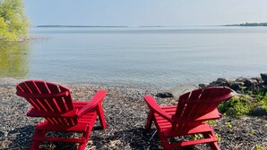 Deux chaises Adirondack rouges sont placées sur une plage de galets, avec un lac paisible et une étendue de terre visible au loin.