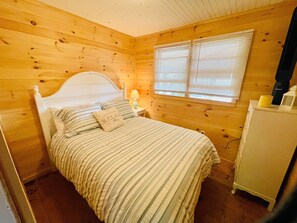 A wooden cabin bedroom with a bed, a nightstand, a lamp, and a window with blinds. - Bright 3 Bedroom Lake Cottage with dock! (North Hero)