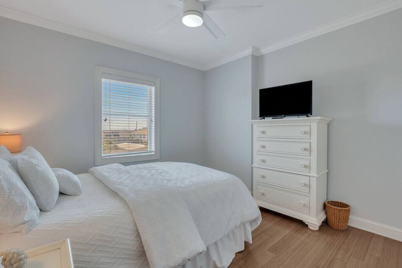 Ein Schlafzimmer mit einem Bett, einer Kommode, einem Fernseher darauf, einem Fenster mit Jalousien und einem Deckenventilator.