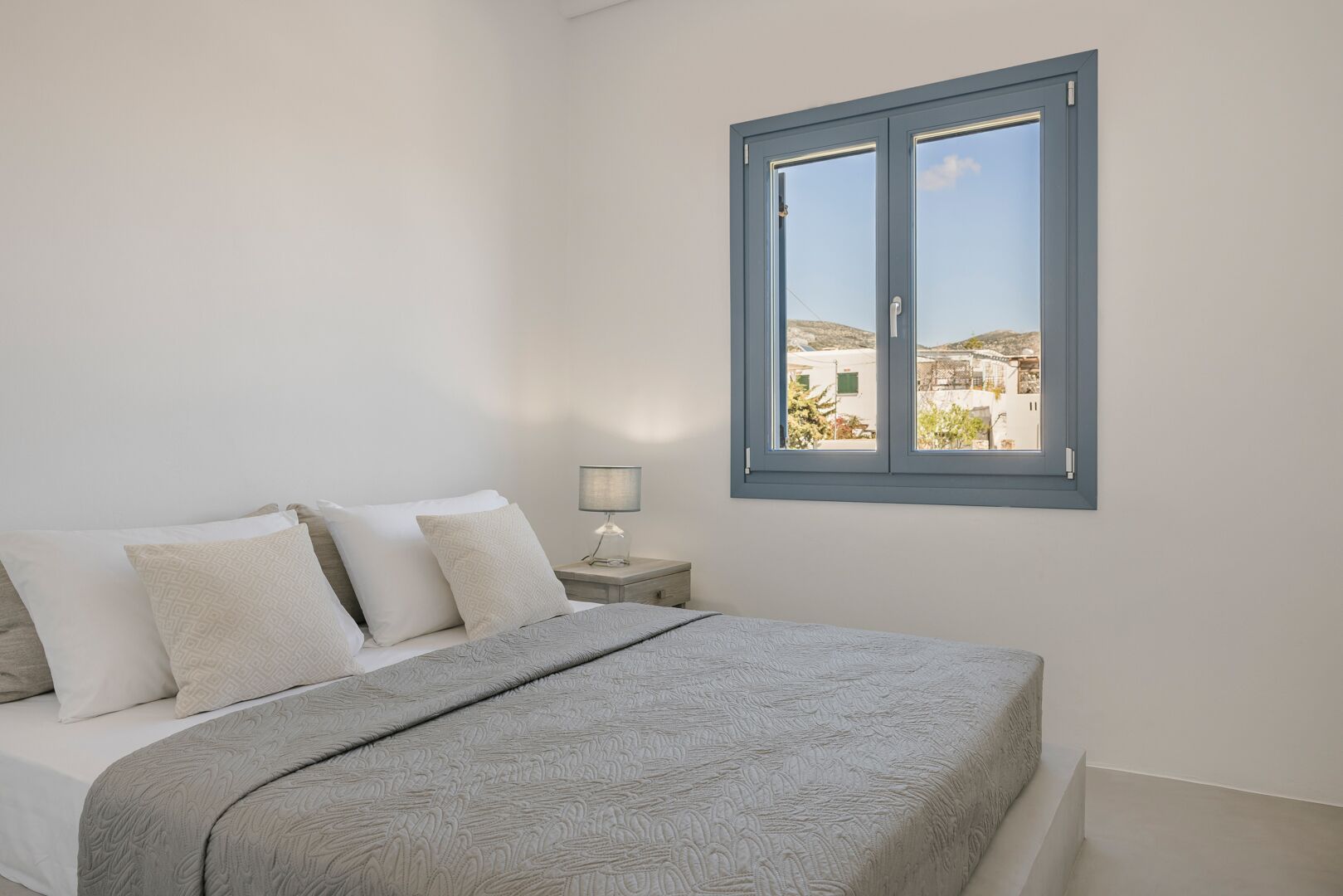 A bedroom with a bed, pillows, a bedside table with a lamp, and a window with a view of buildings.