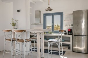 Uma cozinha moderna com armários brancos, uma geladeira de aço inoxidável, uma mesa de jantar com quatro cadeiras e uma janela com vista para o campo.