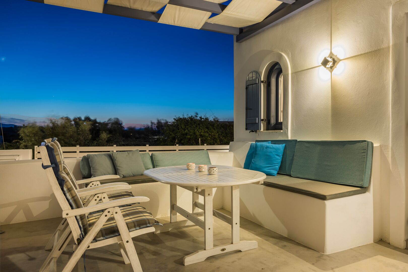 Un espacio al aire libre con mesa y sillas, un banco acolchado y una lámpara fijada a la pared.