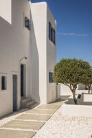 Um edifício branco com uma escadaria que leva a uma porta, uma árvore ao primeiro plano e um céu azul claro.