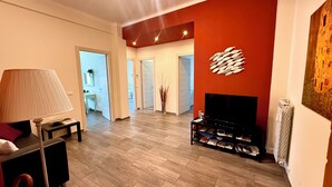 Un saló modern amb televisió de pantalla plana, una taula de centre i una làmpada. Hi ha una estanteria amb revistes i una decoració mural.