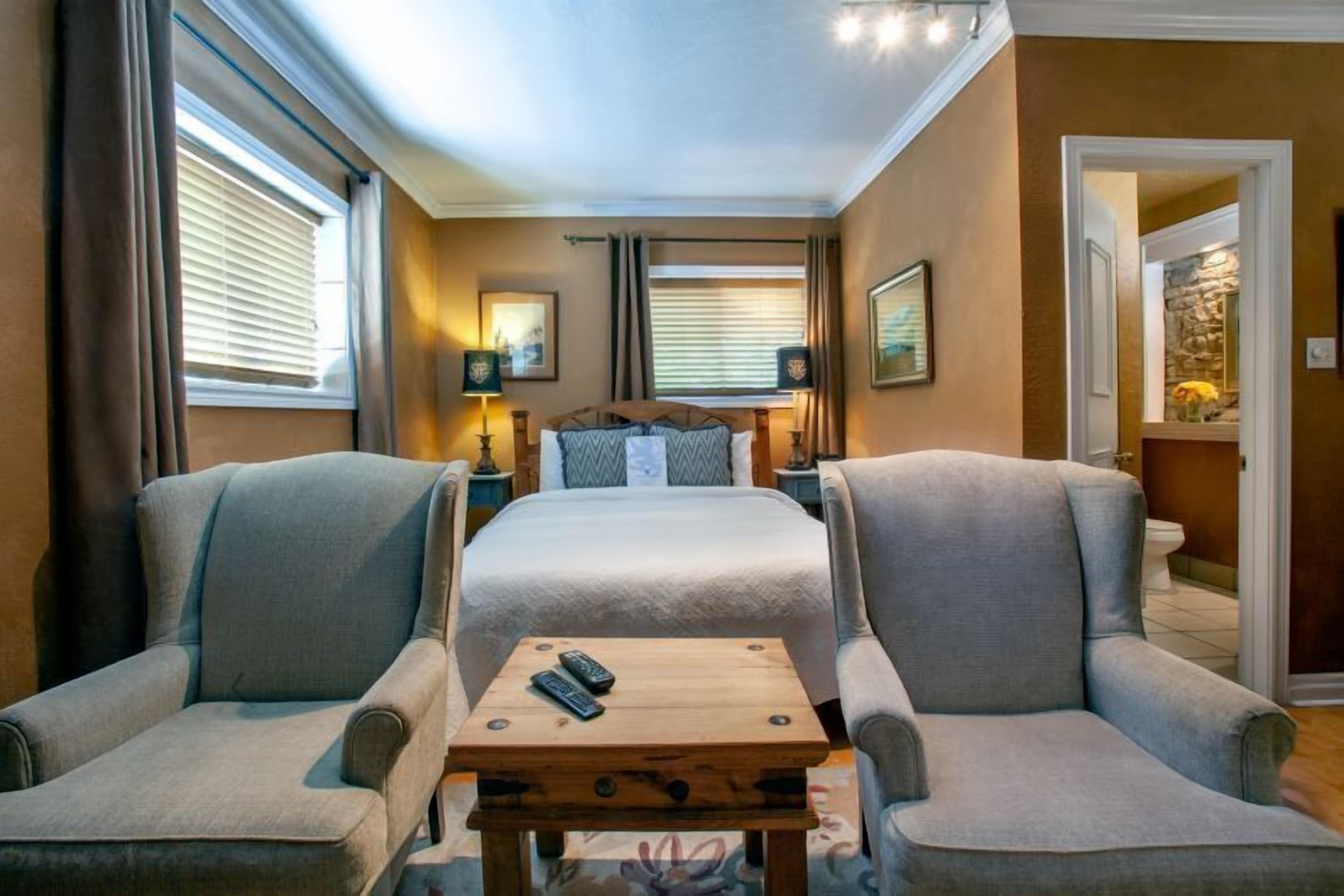 A hotel room with a large bed, two armchairs, a small wooden table, and a nightstand with a lamp.