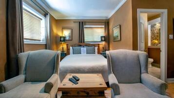 A hotel room with a large bed, two armchairs, a small wooden table, and a nightstand with a lamp.