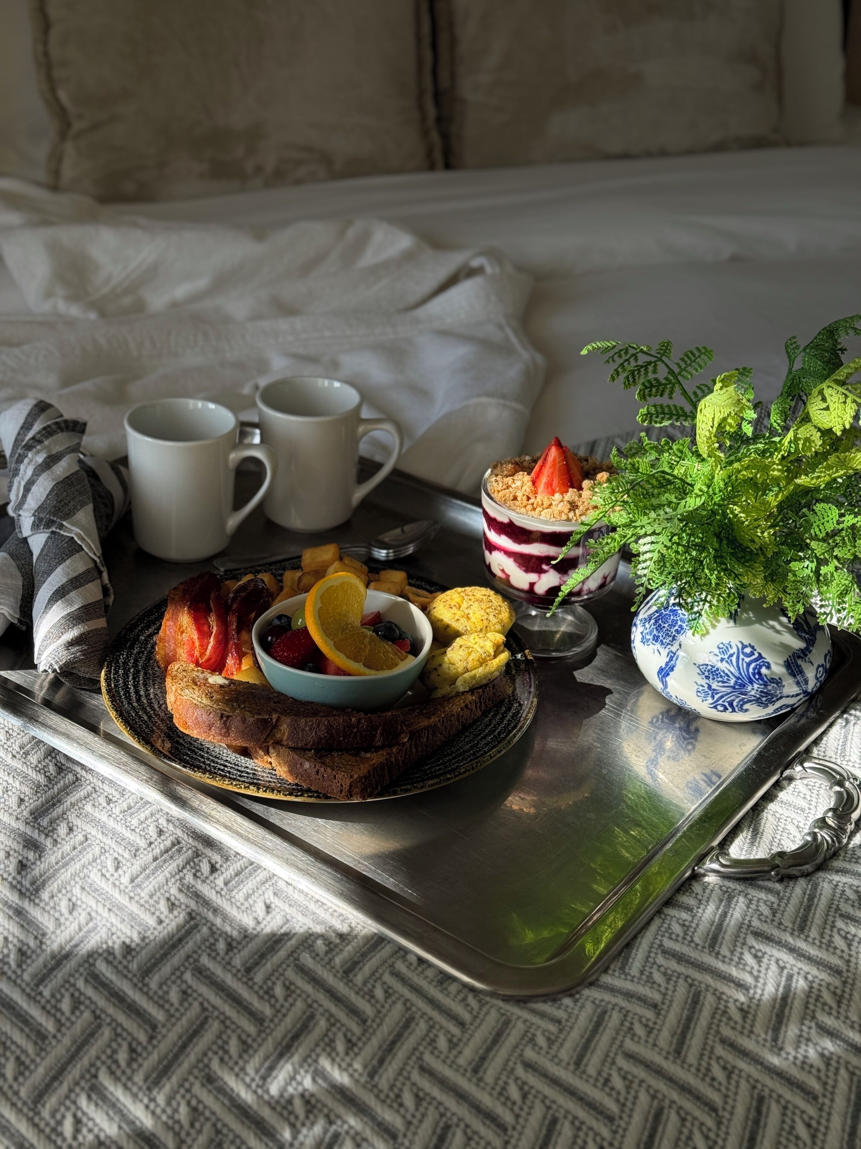 Bandeja de desayuno con tostadas, fruta y un tazón pequeño, junto con dos tazas blancas y una planta decorativa.
