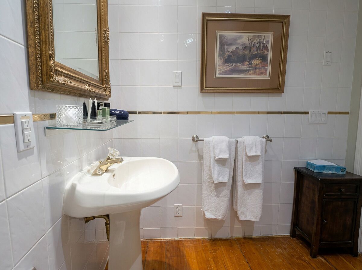 Un baño con lavamanos blanco, espejo con marco dorado, cuadro enmarcado y suelo de madera.