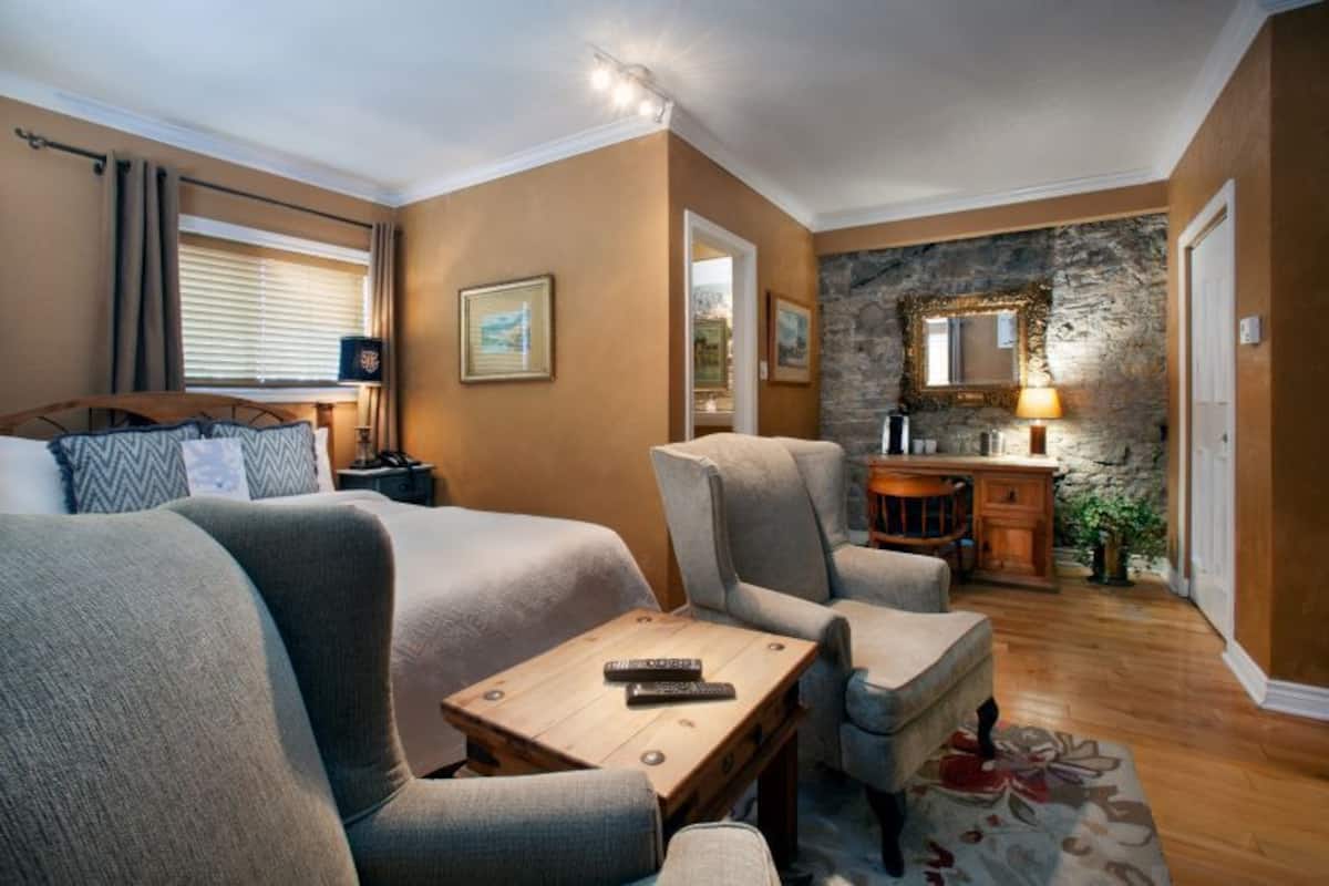 Una habitación con dos camas, una mesita de madera, una silla, una pequeña mesa con lámpara, un espejo y una pared de piedra.