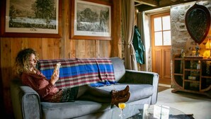 Ein gemütliches Wohnzimmer mit Holzvertäfelung, einem grauen Sofa, einer karierten Decke und einem Glastisch mit einem Getränk.