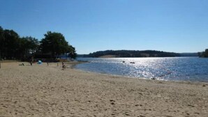 Ein sandiger Strand mit ruhigem Gewässer, Bäumen im Hintergrund und blauem Himmel.
