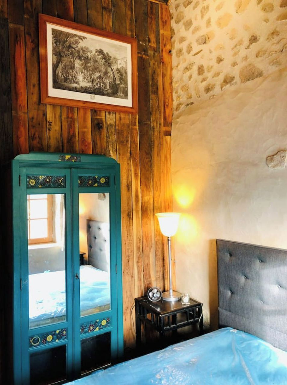 Una habitación con pared de madera, cama con cabecera gris, armario verde azulado, mesita de noche pequeña con lámpara y un cuadro enmarcado en la pared.