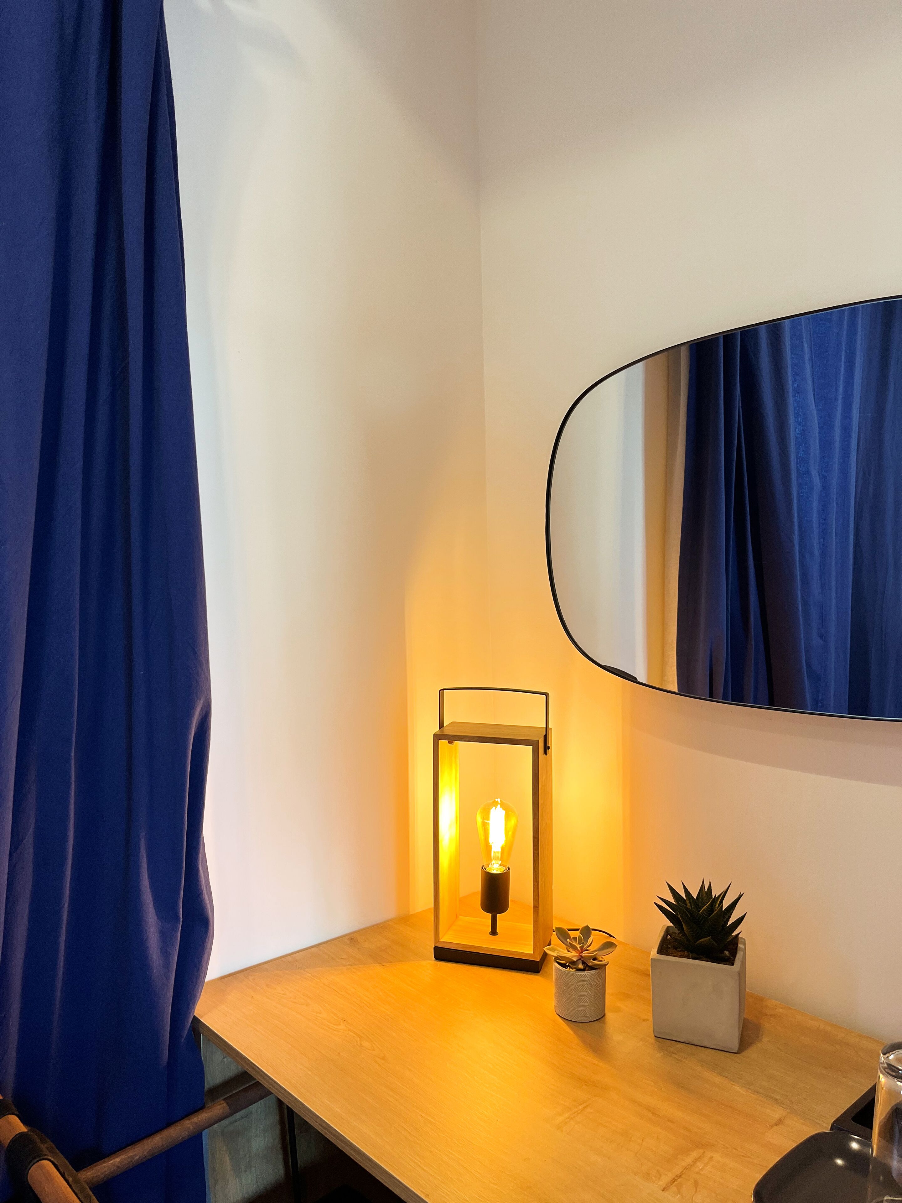 A bathroom with a wooden vanity, a mirror, a blue curtain, a floor lamp, and two potted plants.