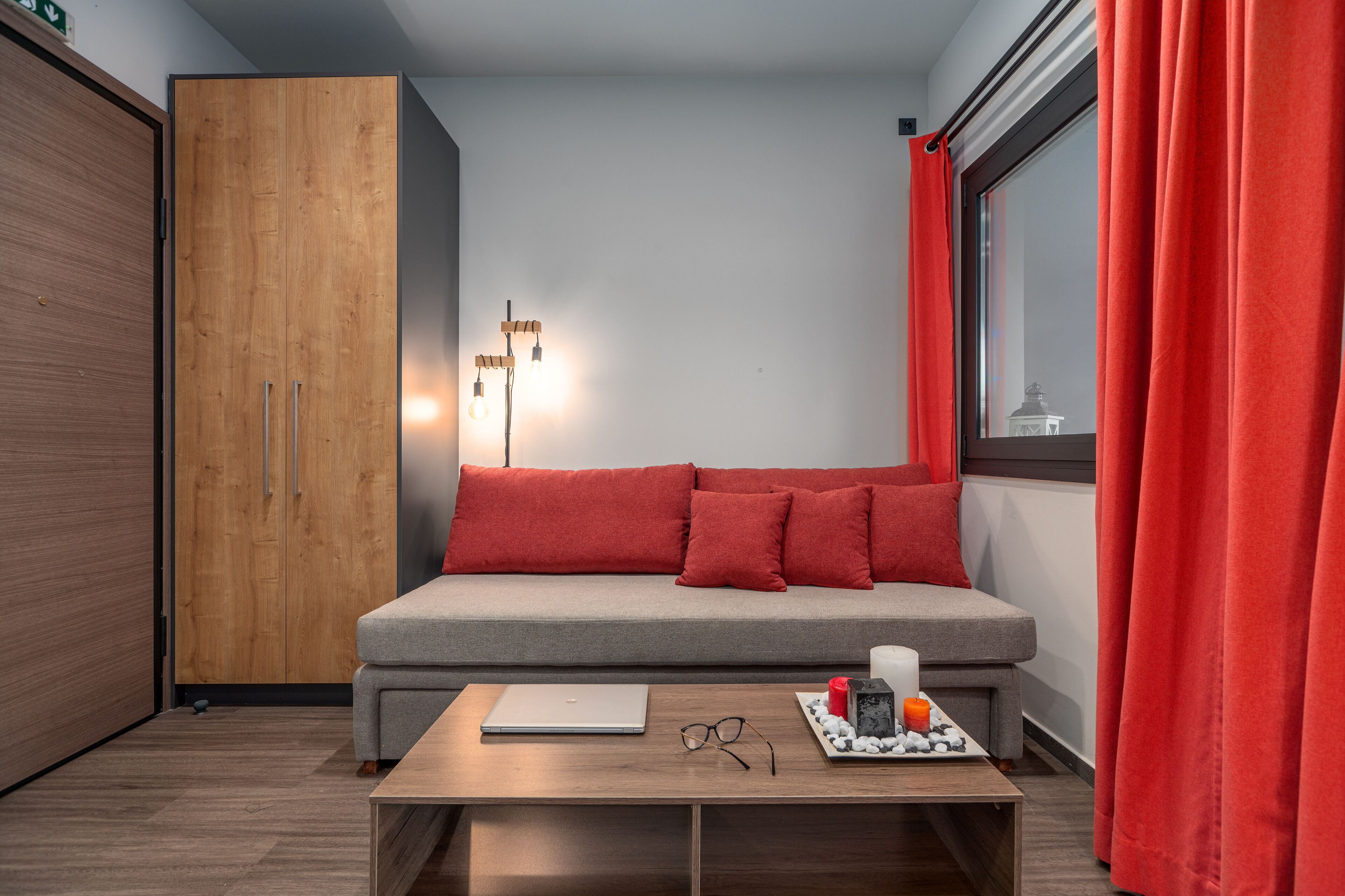 Quarto moderno com sofá cinzento, guarda-roupa de madeira e uma mesa com um laptop, copos e uma vela.
