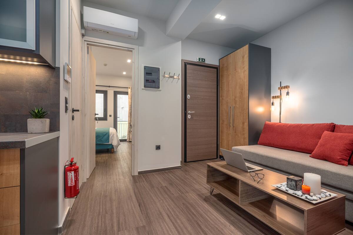 A modern hotel room with a wooden floor, a grey sofa with red cushions, a wooden coffee table with a laptop and decorative items, a wooden wardrobe, and a fire extinguisher on the wall.