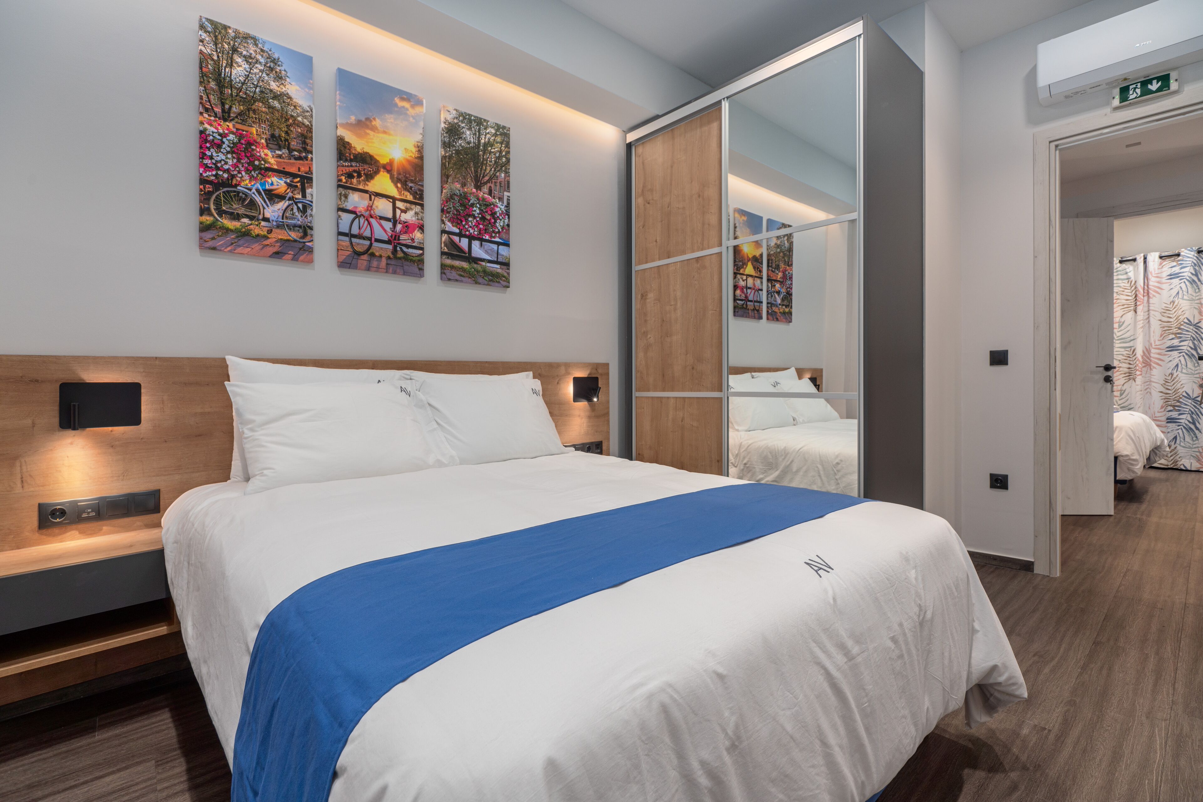 Et moderne hotelværelse med en stor seng, et træhovedgærde og en blå detaljeret løber. Tre indrammede billeder hænger på væggen over sengen.