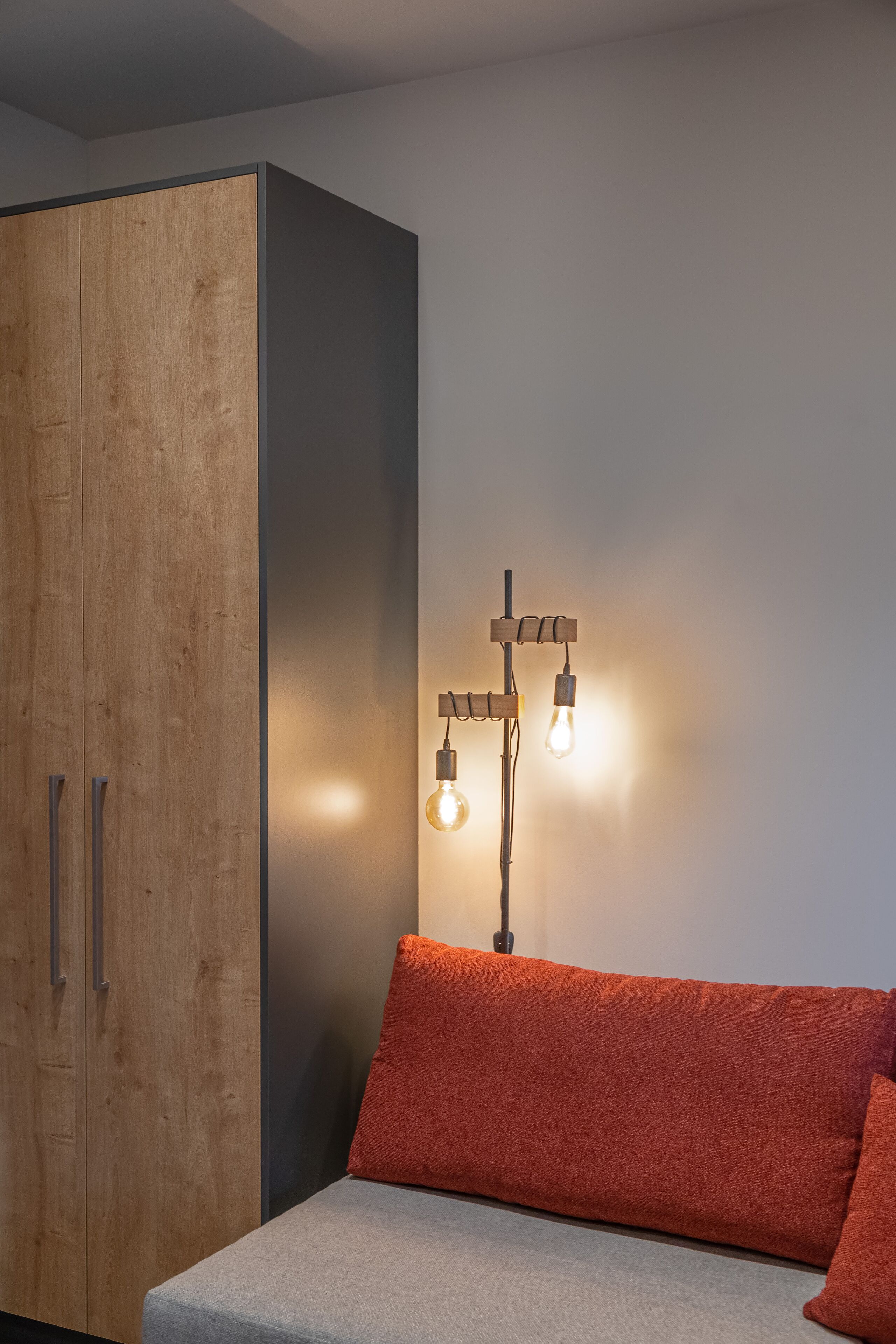Una habitación con un armario de madera, un sofá gris con cojines rojos y una lámpara fijada a la pared.