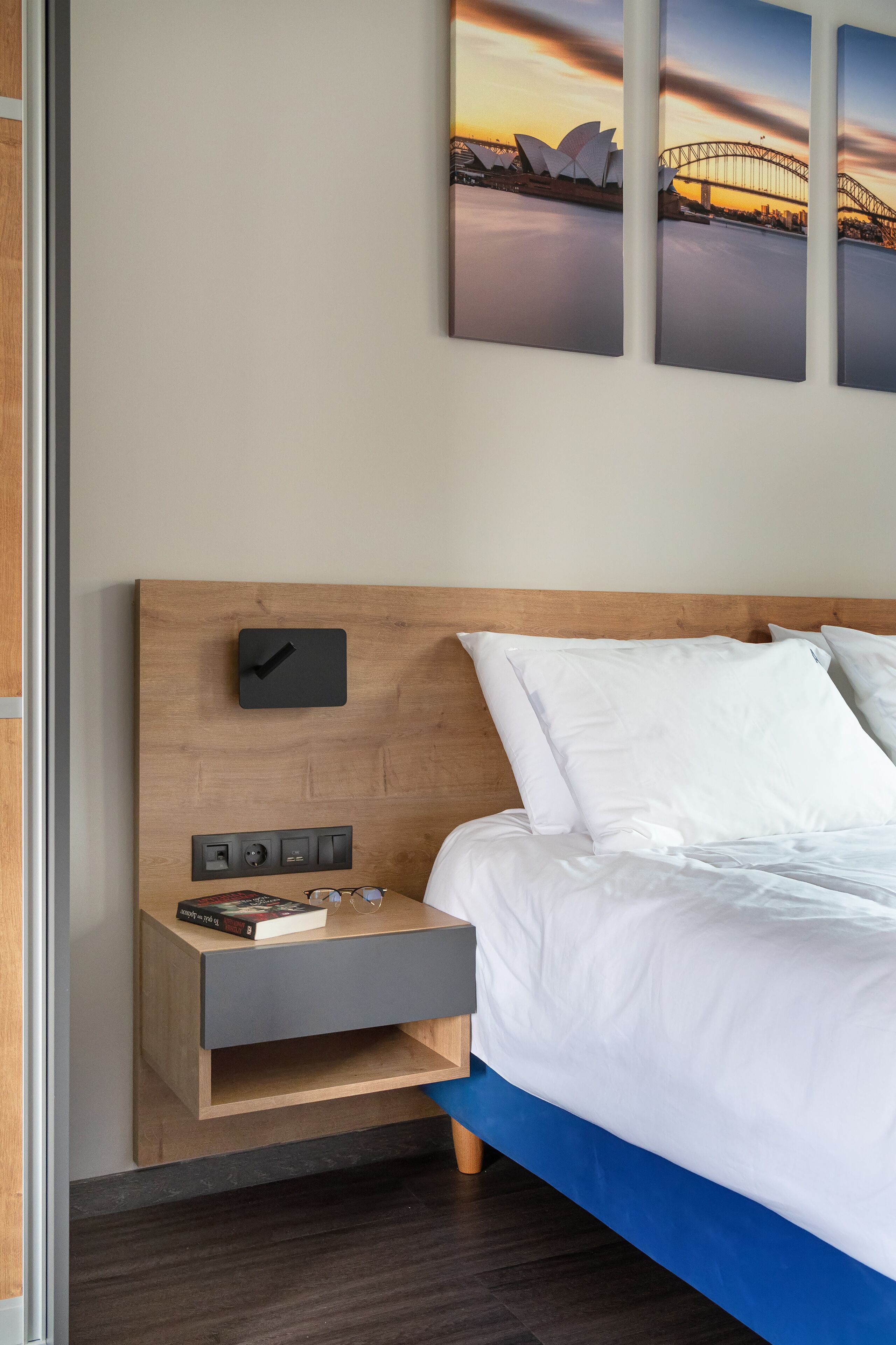 Quarto de hotel moderno com cabeceira de madeira, mesa de cabeceira com relógio, tomada e um livro.