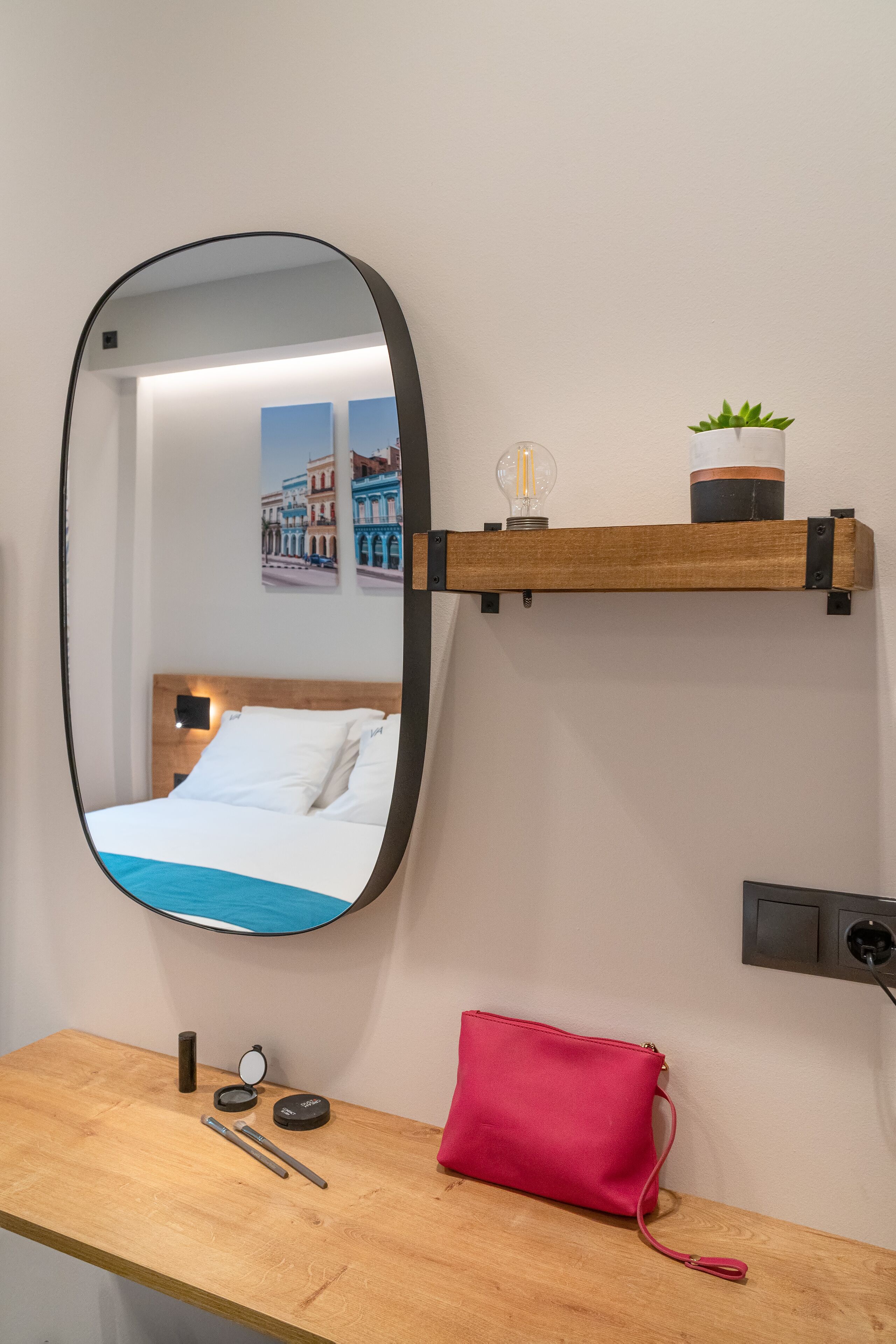 A hotel room with a bed, a wooden nightstand, a mirror, a red handbag, a shelf with a plant and a light bulb, and a wall-mounted power outlet.