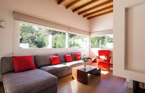 A modern living room with a grey sectional sofa, red cushions, a wooden coffee table, and a red armchair. Large windows provide a view of greenery outside.