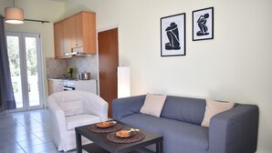 A modern living room with a grey sofa, a white armchair, and a coffee table with two decorative plates. There are framed artworks on the wall and a kitchen area visible through an open door. - Elounda Holidays Argyro (Schisma Elountas)