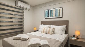 Habitación de hotel con cama, dos lámparas de noche, aire acondicionado de pared y un cuadro abstracto enmarcado.