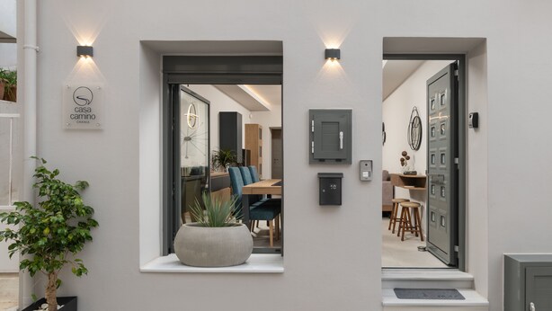 Una entrada moderna con un cartel que dice "casa camino CHANIA", una planta en maceta y una vista al interior que muestra un comedor con sillas y una mesa.