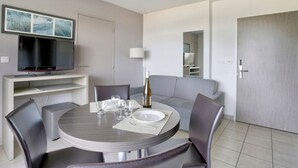 Un espace repas moderne avec une table ronde, des chaises grises, une bouteille de vin et un téléviseur posé sur un meuble.