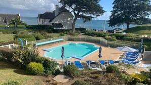 A swimming pool with blue and white lounge chairs, surrounded by a white fence, with a view of the sea and houses in the background.