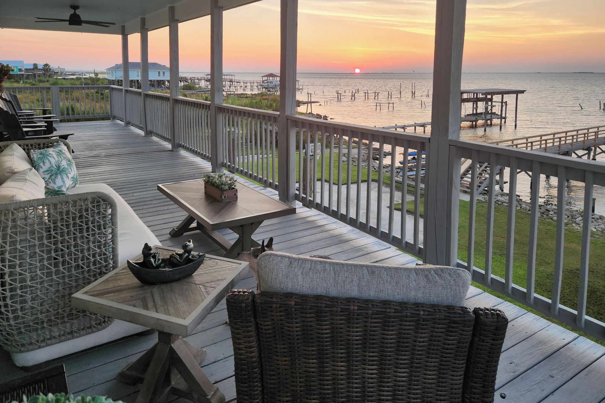 Una terraza techada al aire libre con muebles de mimbre, una mesa con un bol y vistas al atardecer sobre el agua.