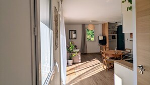 A modern interior with a wooden floor, a dining area with a table and chairs, a kitchen with a TV, and a large window allowing natural light.